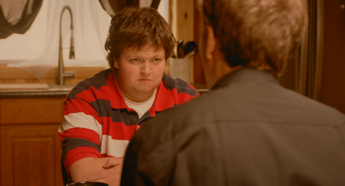 A man in a red and white striped shirt sits at a table facing another person with his arms crossed in a kitchen.