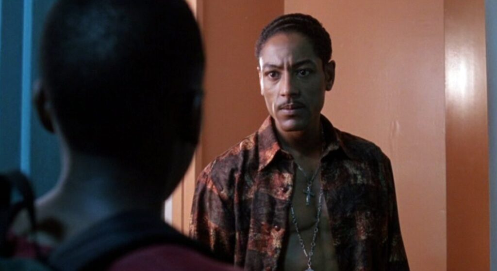 A man wearing a patterned shirt stands in a door frame, facing another person whose back is to the camera. The man's expression is serious.