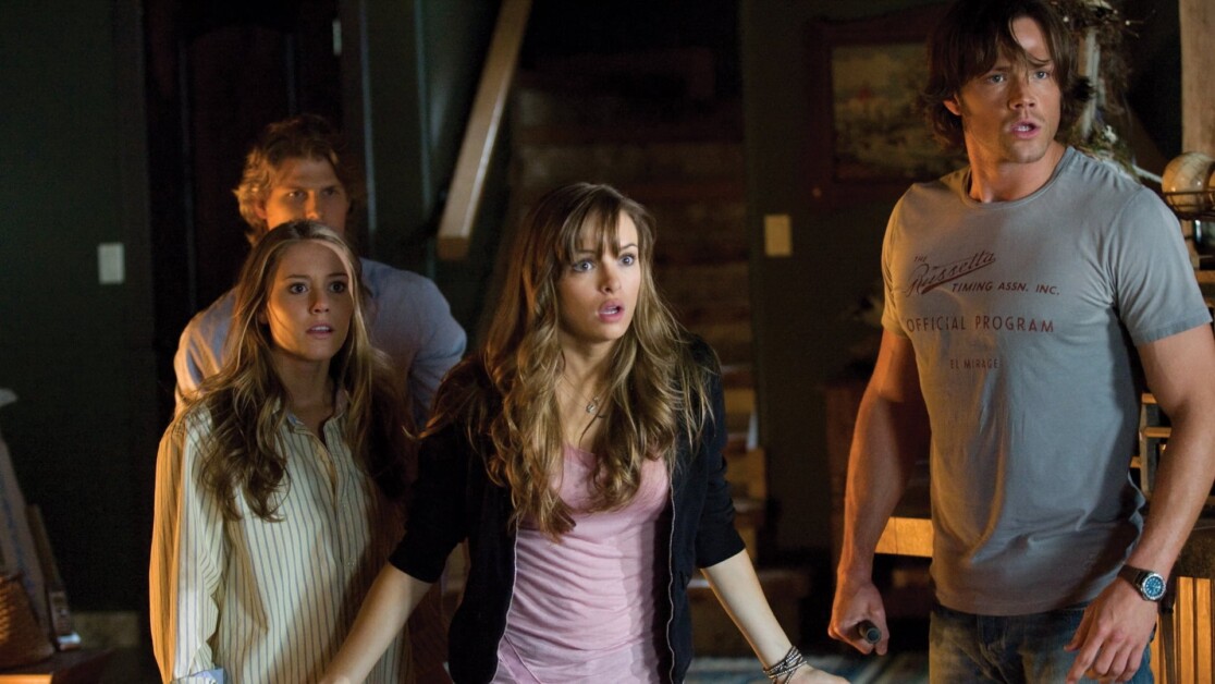 Four people stand closely together in a dimly lit room, looking off to the right with expressions of surprise and concern.