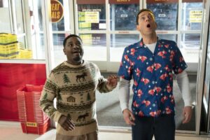 Two men are standing inside a store. The man on the left in a patterned sweater looks worried, while the man on the right in a colorful shirt seems surprised. Bright baskets and a glass door are behind them.