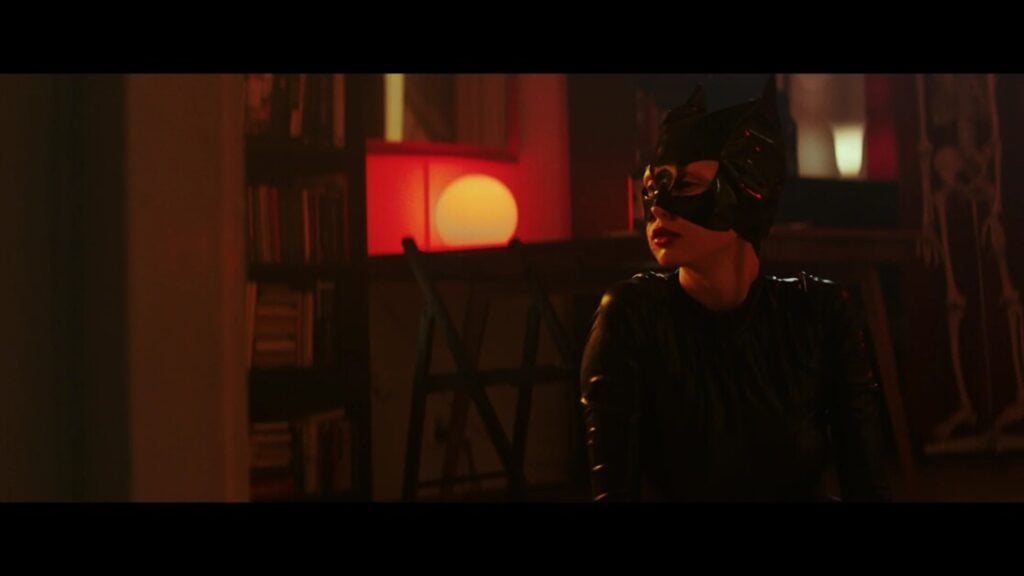 Person in a black cat costume with a mask and ears, sitting in a dimly lit room with a red-lit lamp and bookshelves in the background.