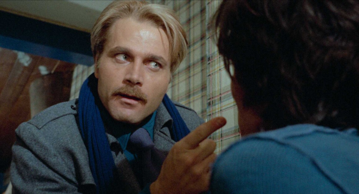 A man with a mustache and light hair points his finger while talking to another person who is facing away.