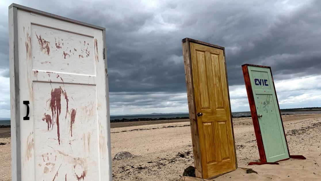 Three standing doors on a sandy beach under a cloudy sky. The left door is white with brown markings, the middle door is wooden and plain, and the right door is green with "EVIE" written on it.