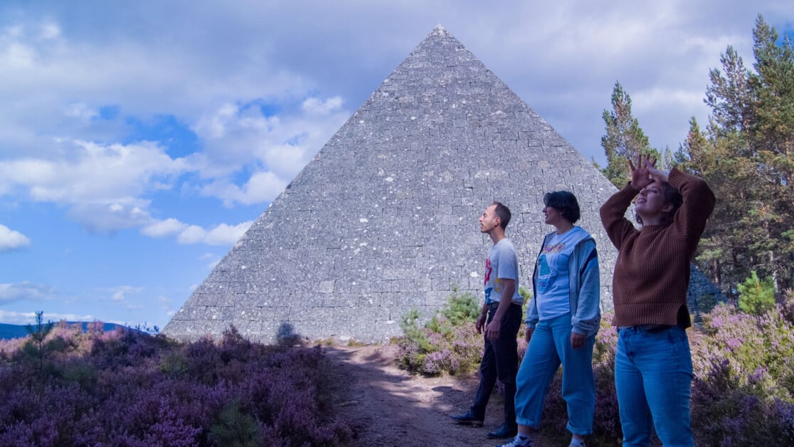 Three people stand on a path with a large stone pyramid in the background. One person is shielding their eyes, looking up. The scene includes bushes and trees under a partly cloudy sky.