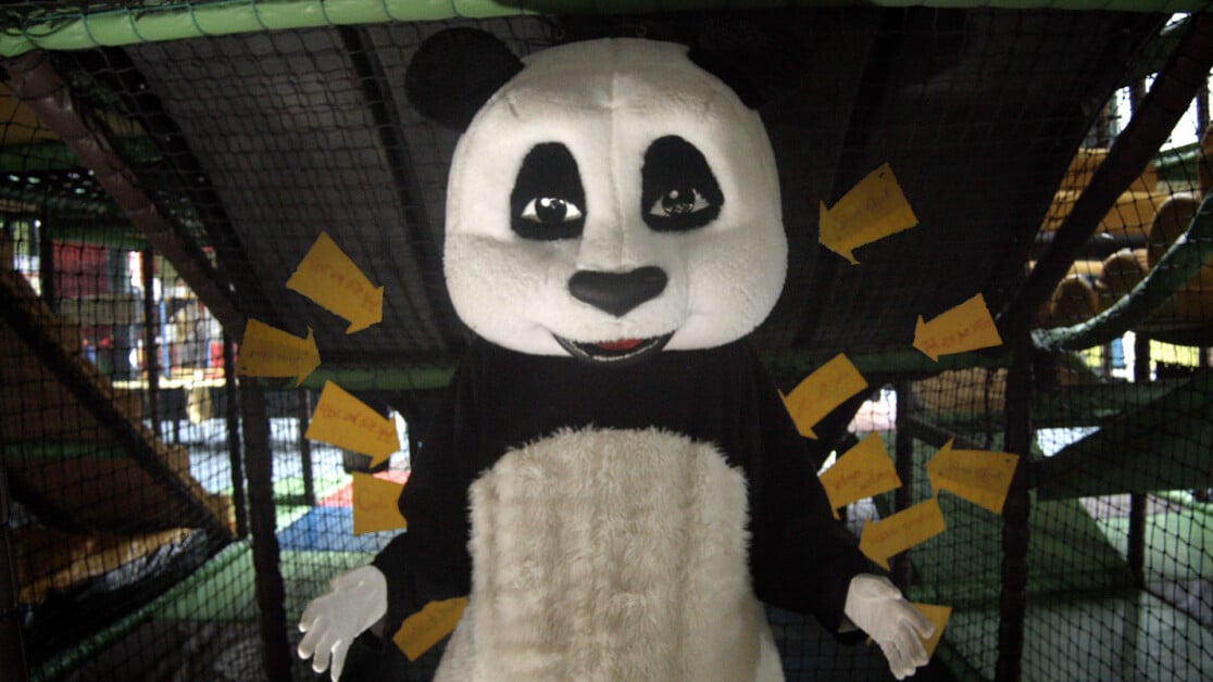 Person wearing a panda costume stands in front of a black net with yellow papers attached to it.
