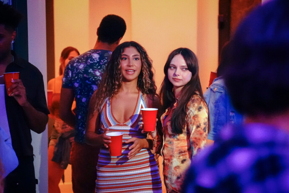 Two women stand together at a party, each holding a red cup. One woman smiles and wears a striped dress, while the other, in a patterned dress, looks off to the side.