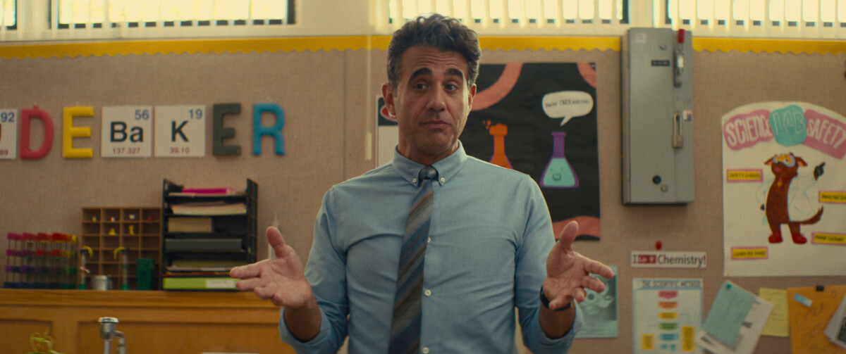 A man in a blue shirt and tie stands in a classroom, gesturing with his hands. The background has educational posters, including chemistry-related items and colorful alphabet letters.