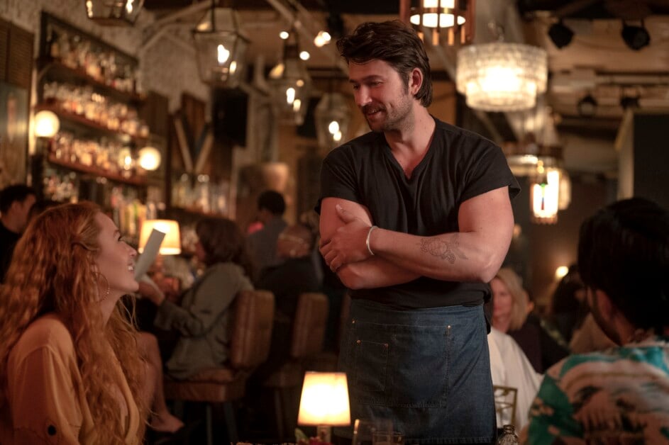 A person with long hair sits at a table in a dimly lit restaurant, talking to a standing waiter who is wearing a black shirt and apron. Other diners and bar shelves are visible in the background.