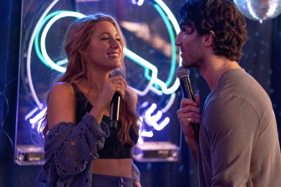 Two people hold microphones while singing together in front of a neon-lit backdrop. Both appear to be enjoying the moment.