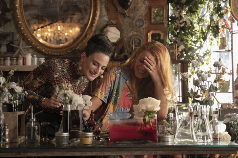Two women smile and chat while working in a cluttered floral workshop filled with various plants, flowers, and glass containers. One woman touches her head, and both seem engaged in a light-hearted moment.