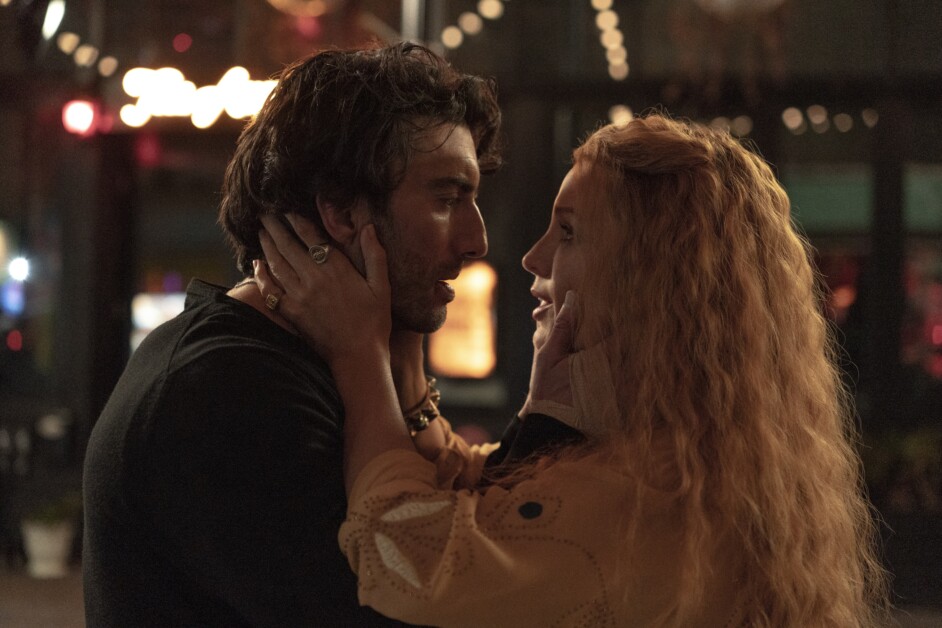 A man and a woman stand closely, with the woman holding the man's face in her hands, looking into each other's eyes. They appear to be having an intimate moment.