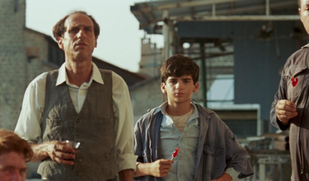 An older man and a young boy, both in casual attire, stand outdoors. The boy holds a red object. The background features industrial structures.