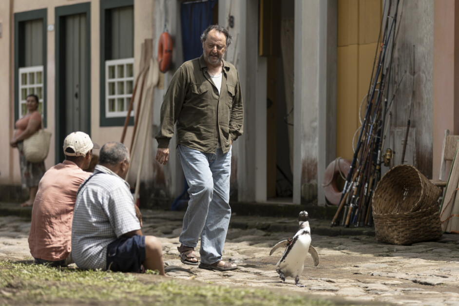 A man walks alongside a penguin on a cobblestone path as two seated men observe them. Fishing rods and a basket are visible in the background near a building.