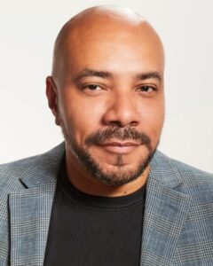A bald man with a beard, wearing a black shirt and a gray plaid blazer, looks at the camera with a neutral expression against a plain background.