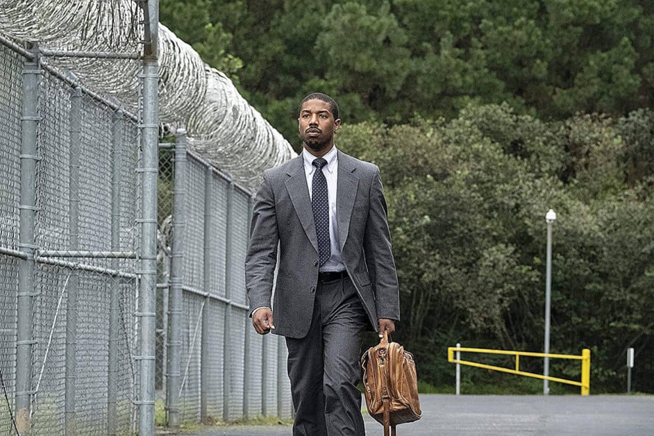 A man in a gray suit and tie walks beside a chain-link fence topped with barbed wire, holding a brown leather bag, with trees visible in the background.