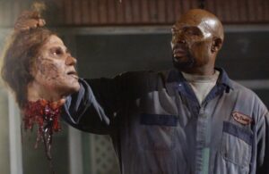 A man holds a severed, bloody, and decayed head in his right hand. He is wearing a dirty blue jumpsuit and appears serious.