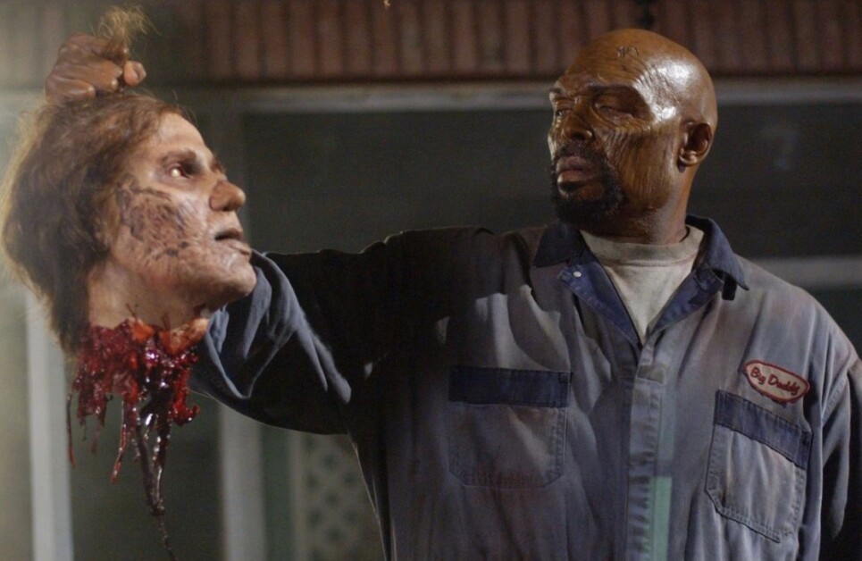 A man holds a severed, bloody, and decayed head in his right hand. He is wearing a dirty blue jumpsuit and appears serious.