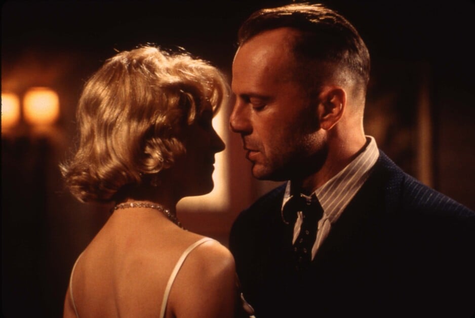 A man in a suit with a shaved head and a woman with blonde hair wearing a white dress are standing close together, looking at each other in a dimly lit setting.