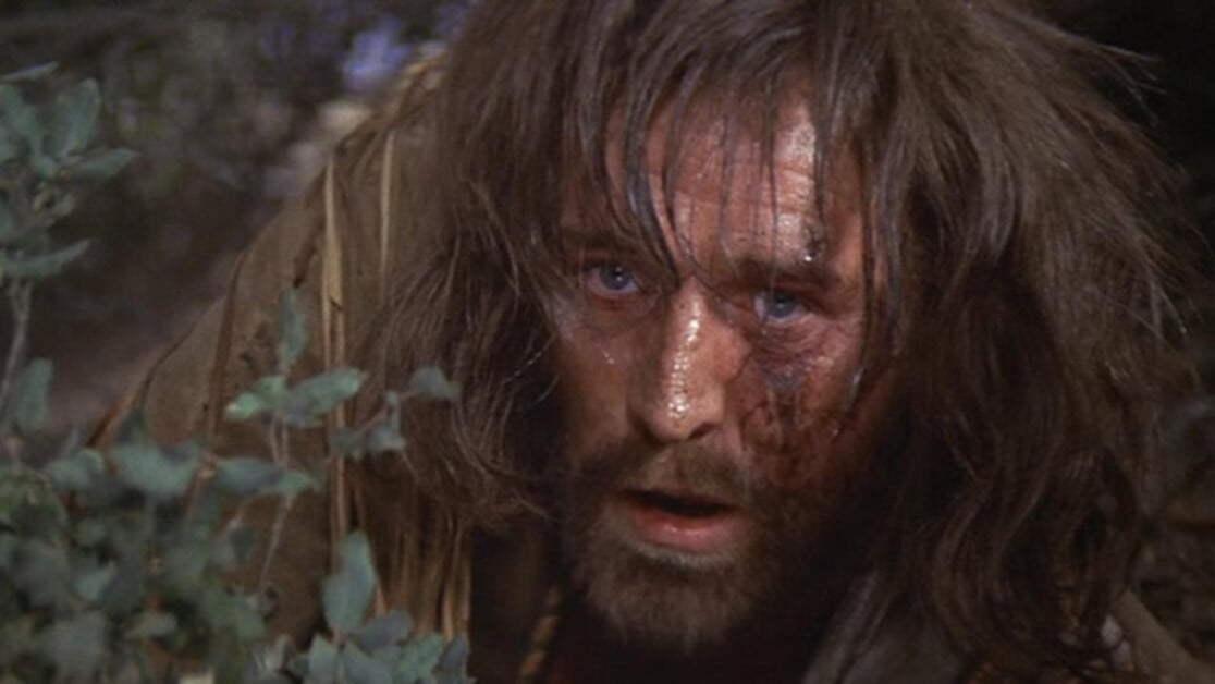 A bearded man with long hair and a dirt-smeared face is hunched down among foliage, looking up with a serious expression.