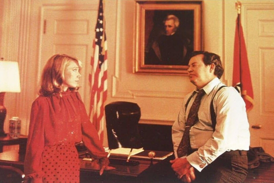 A woman in a red dress stands next to a man in a shirt and tie who is leaning against a desk in an office with a U.S. flag, another flag, and a portrait on the wall.