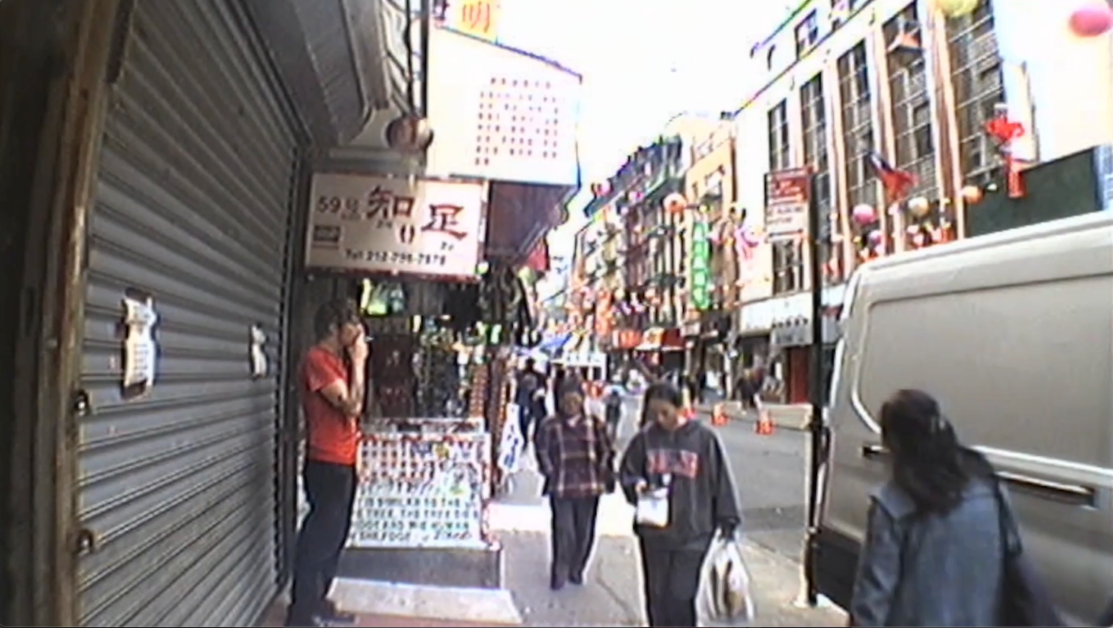 People walk down a bustling street lined with shops and colorful decorations. A person stands by a storefront, speaking on the phone. A white van is parked on the right side of the image.