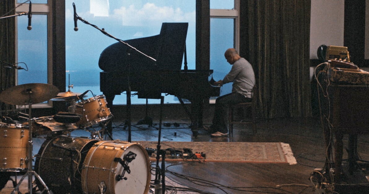A man plays a grand piano in a room with drums and other recording equipment, next to large windows showing a view of the sky.