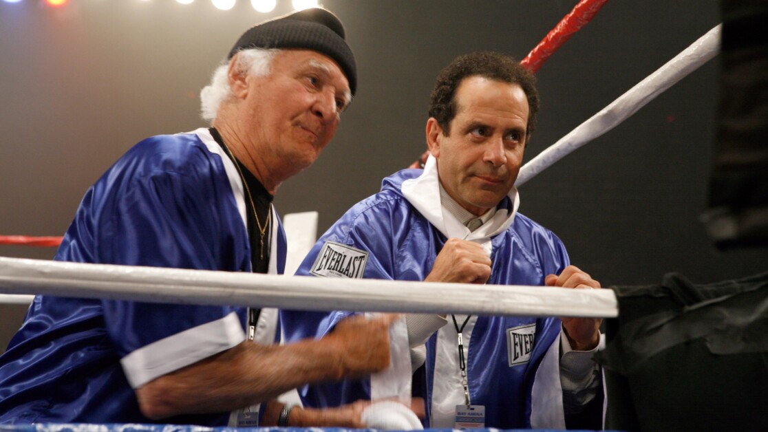 Two men in blue Everlast boxing robes are sitting in a boxing ring. One is older with a white beard and wearing a black beanie, while the other has dark hair and holds a white towel.