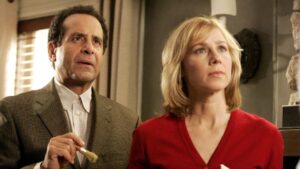 A man holding a brush stands beside a woman in a red top, both looking off-camera with concerned expressions. They are in an indoor setting with a curtain and a wall lamp in the background.