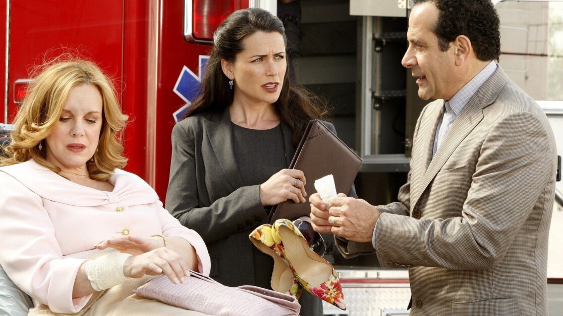 Three people are conversing in front of an ambulance; a woman seated in a pink jacket, a standing woman holding documents, and a man in a suit holding paper.