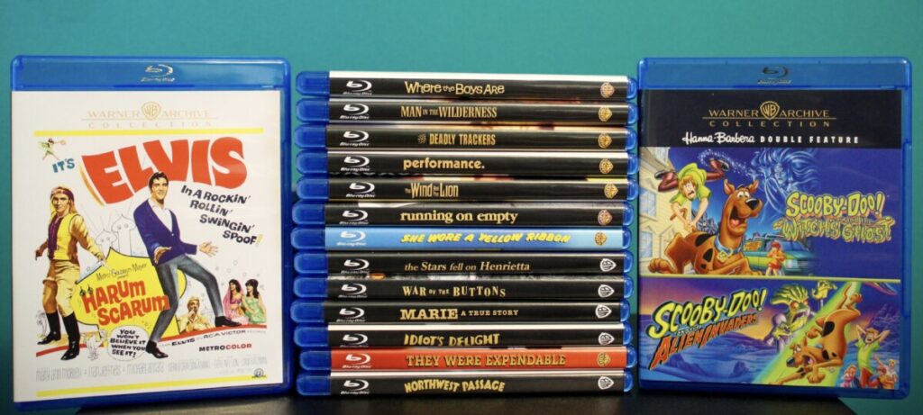 A collection of Blu-ray films displayed upright, featuring titles such as "It's Elvis," "When Dinosaurs Ruled the Earth," "Scooby-Doo Where Are You!," "Scooby-Doo and the Reluctant Werewolf," and others.
