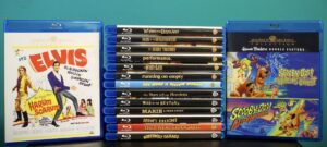 A collection of Blu-ray films displayed upright, featuring titles such as "It's Elvis," "When Dinosaurs Ruled the Earth," "Scooby-Doo Where Are You!," "Scooby-Doo and the Reluctant Werewolf," and others.