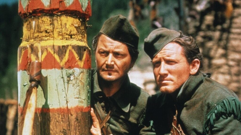 Two men dressed in green medieval attire are hiding behind a decorated wooden pole, peering ahead with focused expressions.