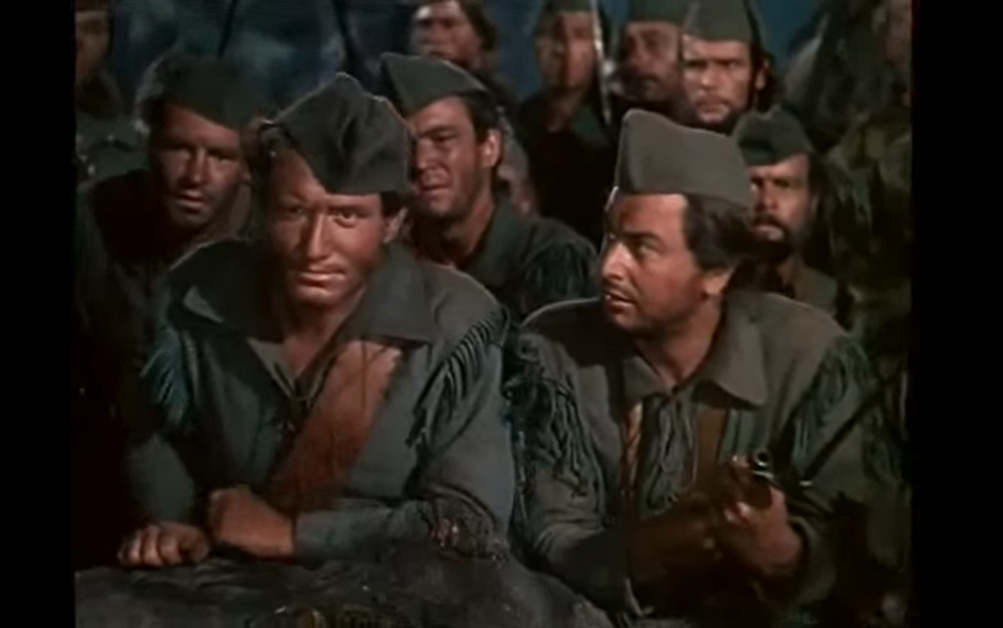 A group of men in matching green military attire and hats are seated closely together, looking ahead with serious expressions. Some men in the background hold rifles.