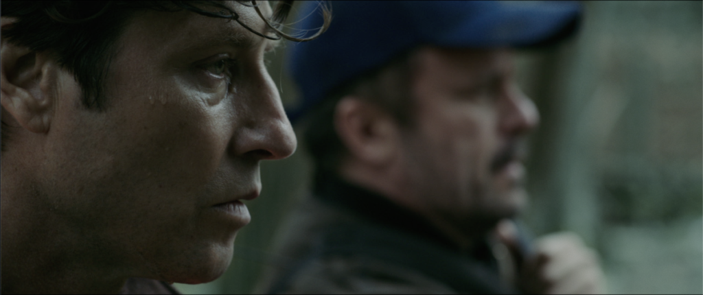 Two men appear in the foreground, one in profile with a focused expression, and another in a blue cap in the background, both seemingly outdoors in a blurred setting.