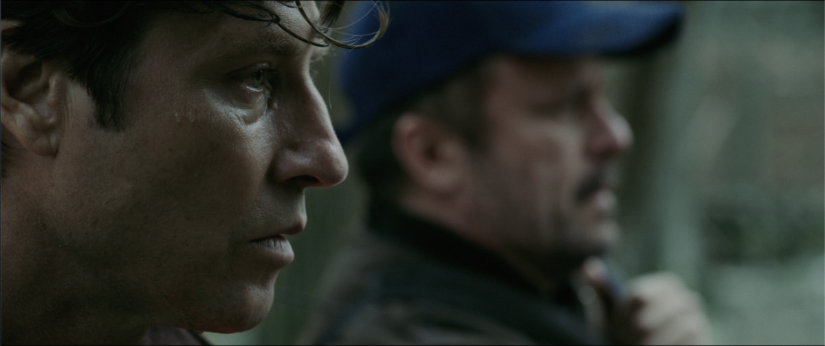 Two men appear in the foreground, one in profile with a focused expression, and another in a blue cap in the background, both seemingly outdoors in a blurred setting.