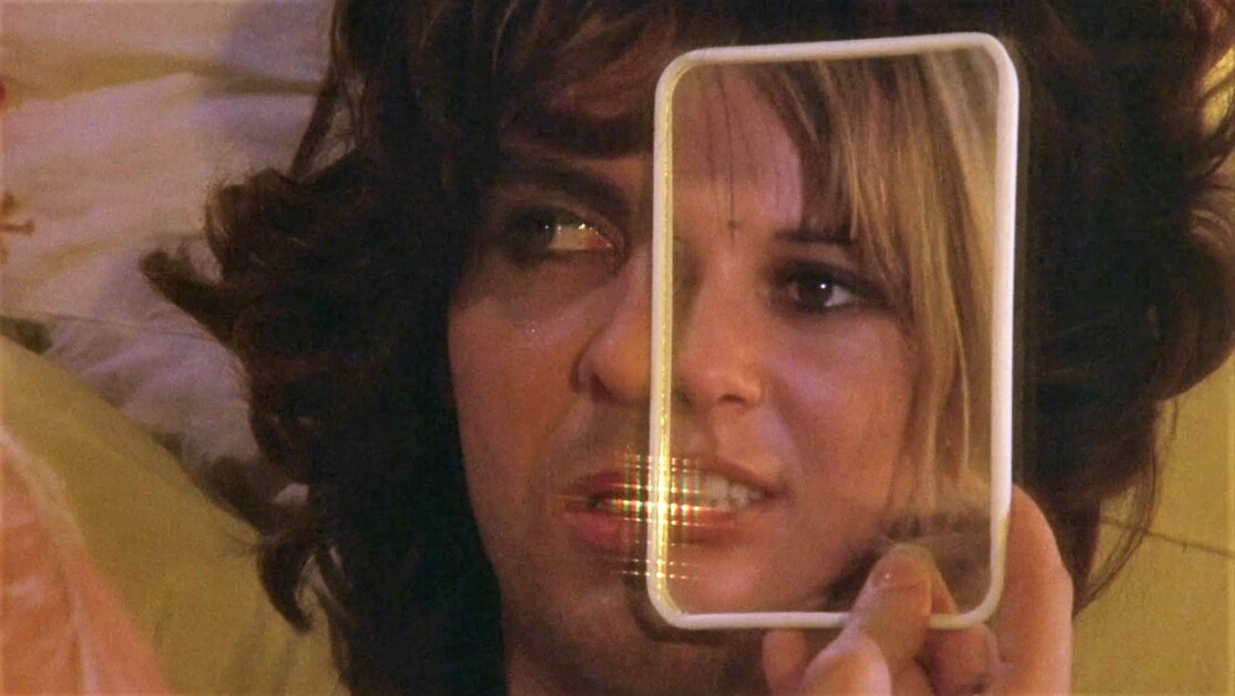A person holds a small rectangular mirror in front of their face, reflecting half of their face with an expression that appears contemplative. The background is out of focus.