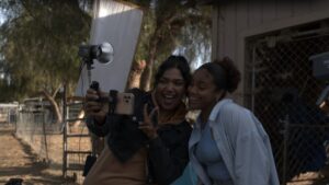 Two women standing outside, smiling and taking a selfie with a smartphone on a sunny day. Trees and a building are visible in the background.