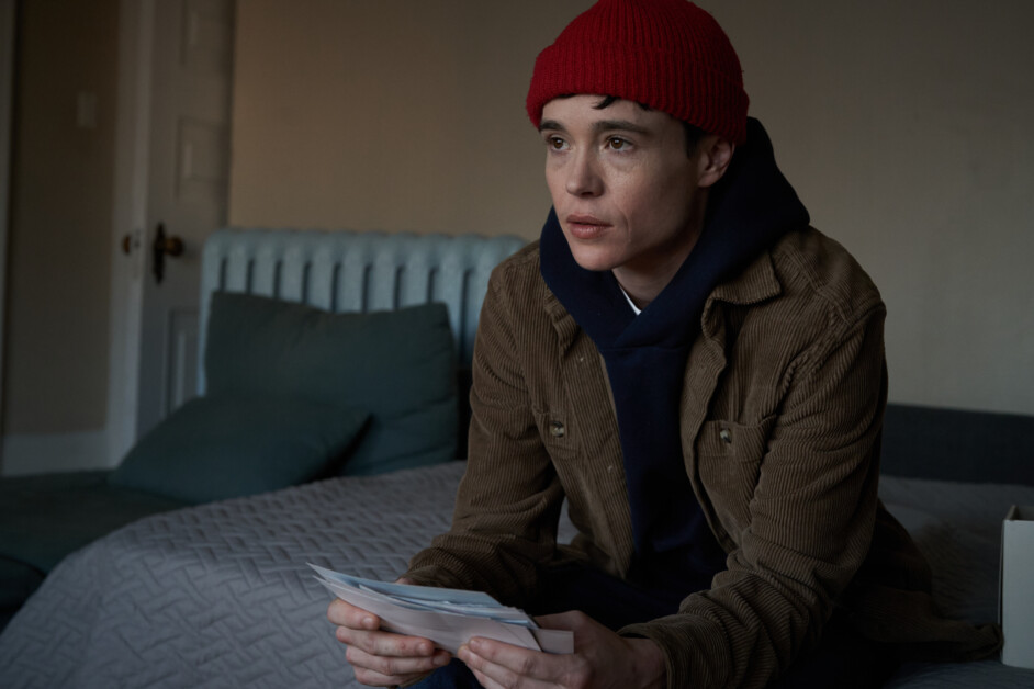 A person wearing a red beanie and brown jacket is sitting on a bed holding a stack of papers, with a serious expression on their face.