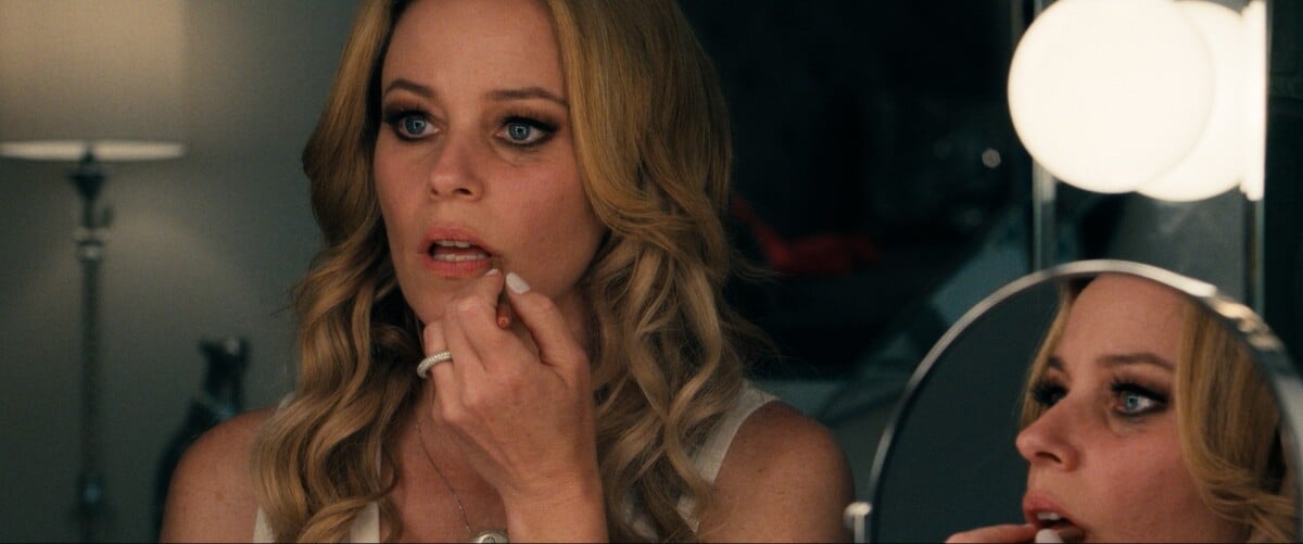 A woman with blonde hair applies lipstick while looking into a mirror under warm lighting.