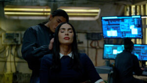 A person sits with eyes closed in a dimly lit room as another person stands behind, touching their head. Computer monitors with blue screens are visible in the background.