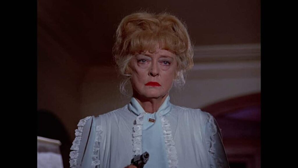 An older woman with red lipstick and a serious expression holds a gun while wearing a light blue outfit with lace details.