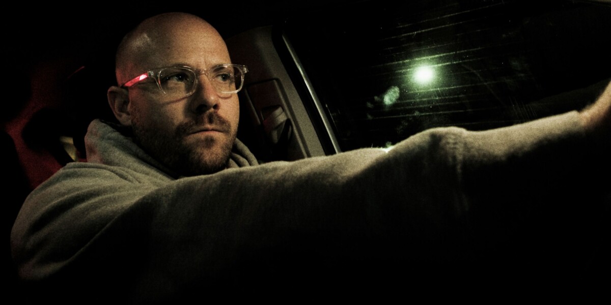 A person wearing glasses and a hoodie is seen driving at night, with a focused expression and one hand on the steering wheel.