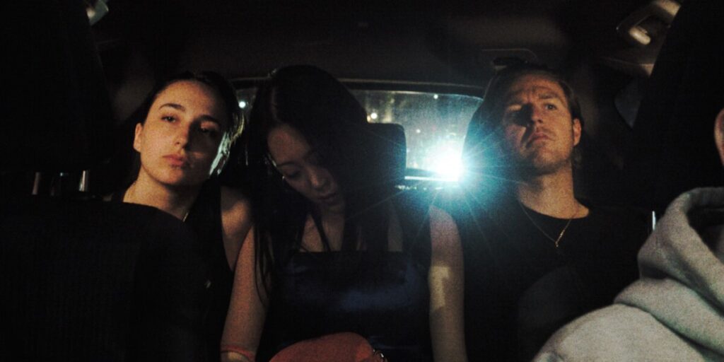 Three people are seated in the back of a car at night, illuminated by dim lighting, with a bright light source behind them.