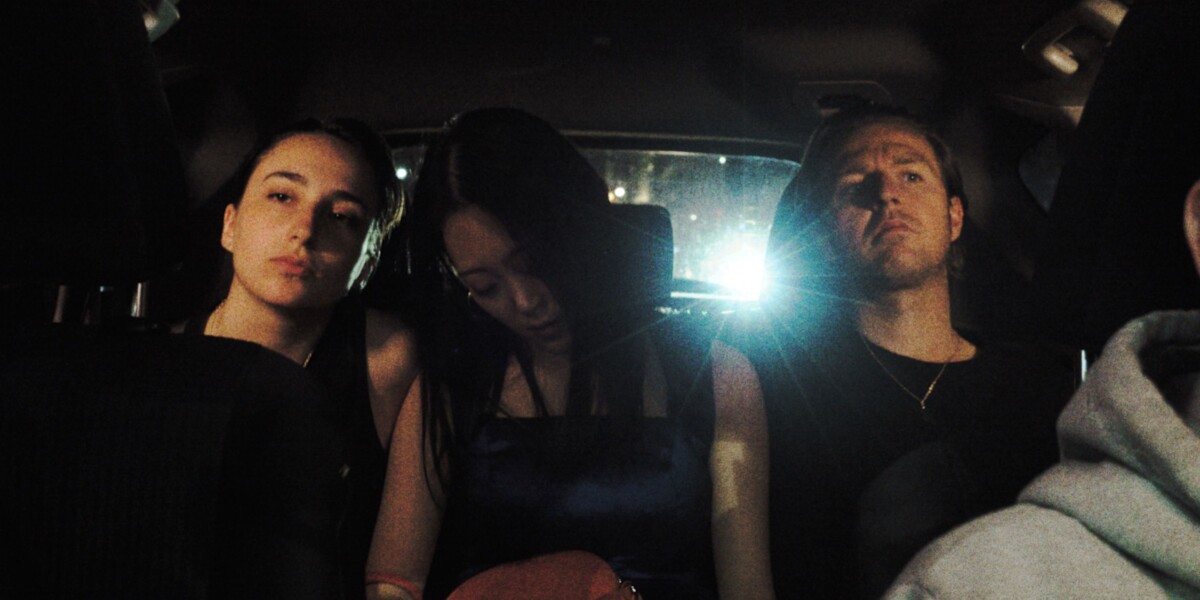 Three people are seated in the back of a car at night, illuminated by dim lighting, with a bright light source behind them.