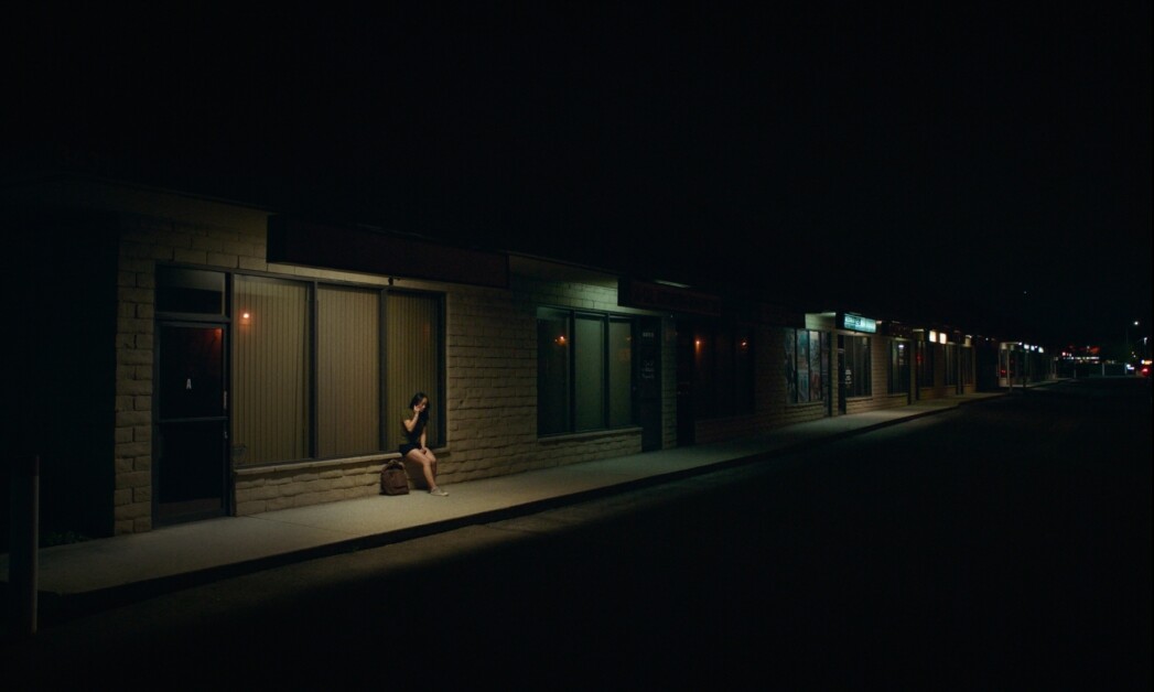 A person sits alone on an outdoor bench in front of a dimly lit storefront at night.