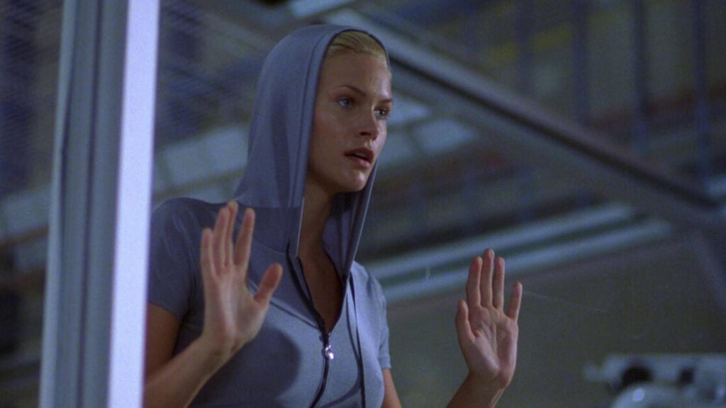 A person in a hooded jacket stands behind glass with hands raised, looking outward with a concerned expression.