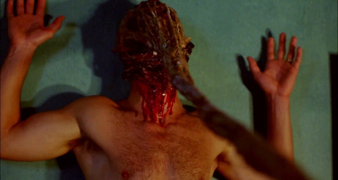 A shirtless person with a disfigured, bloody face is seen laying on their back against a wall, with hands raised.