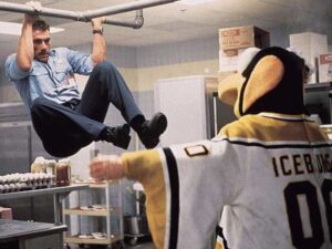 A man in a security uniform performs a pull-up on a metal pipe in a room, while a person in a penguin costume and hockey jersey labeled "ICEBURGH 00" looks up at him.