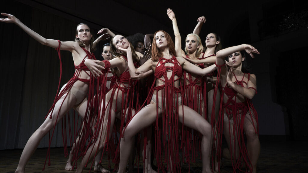 A group of dancers in red costumes striking dramatic poses with expressive arm movements on a dimly lit stage.