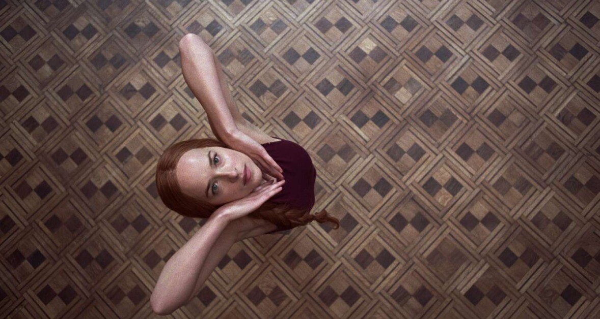 A person with red hair wearing a burgundy outfit looks up with hands framing the face, standing on a patterned wooden floor.